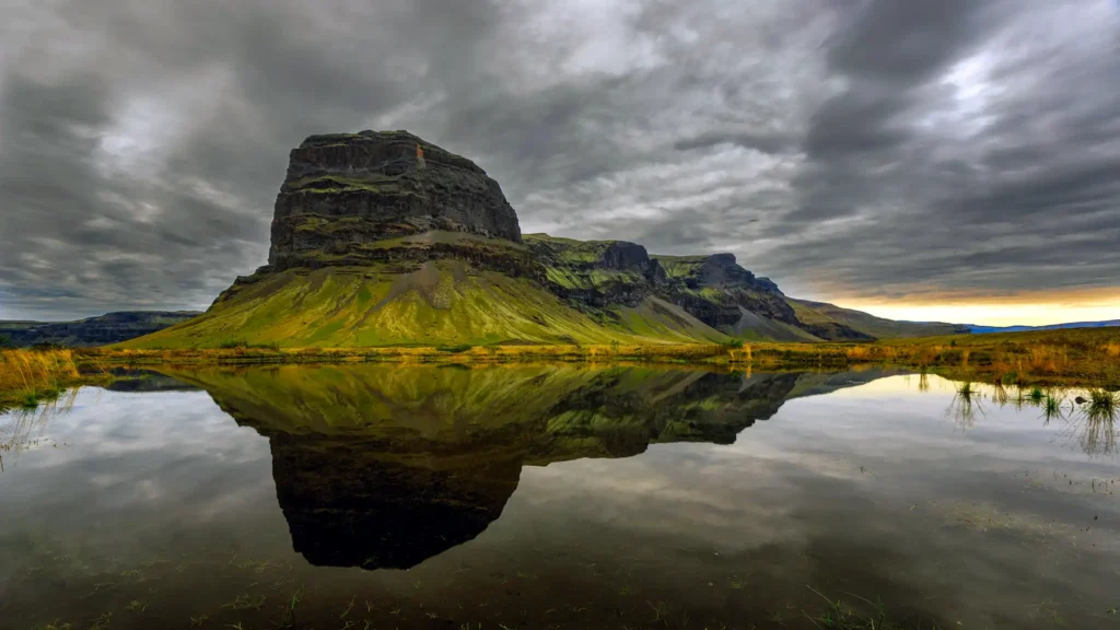 Lómagnúpur volcanic cliffs rising from a moss-covered plain, reflected in still water beneath a dramatic Icelandic sky.
