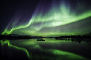 The northern lights sweeping across a clear Icelandic night sky, reflected in still water beneath distant hills.